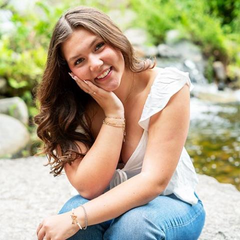 Smiling student sits outdoors on a rock, hand on cheek, with green foliage and a stream.